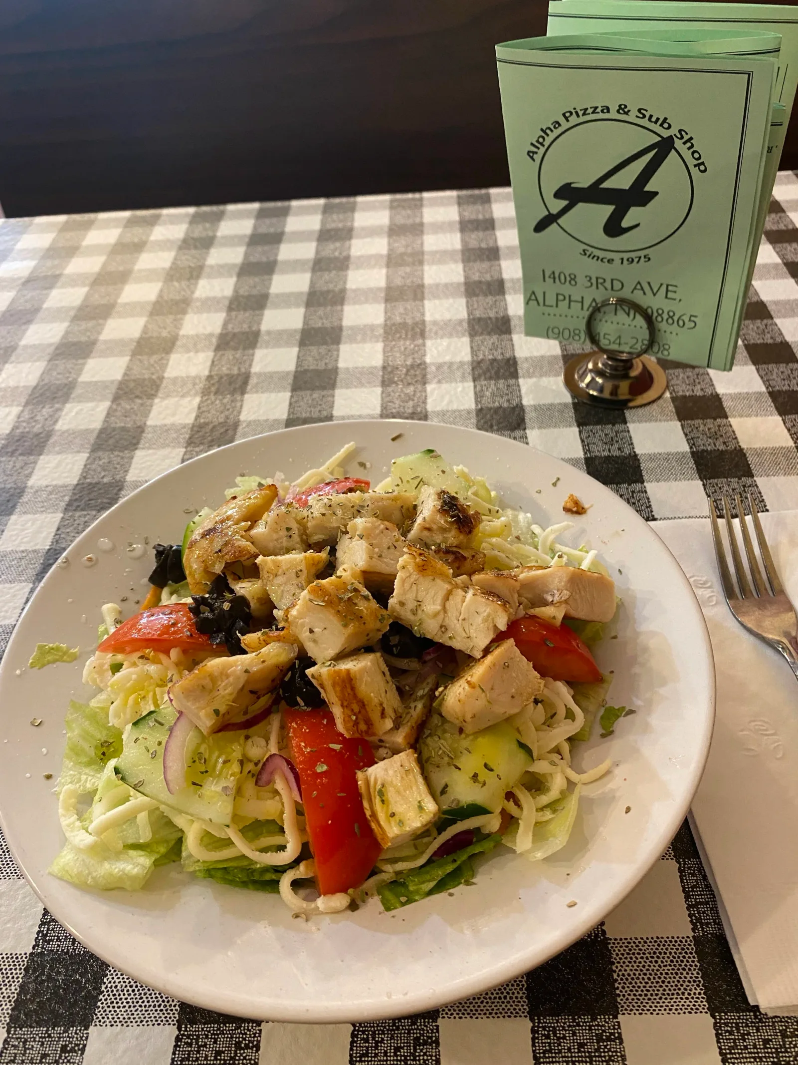 A plate of salad with grilled chicken, lettuce, tomato, cucumber, and red onion, on a checkered tablecloth, with a fork, napkin, and green paper menu in the background.